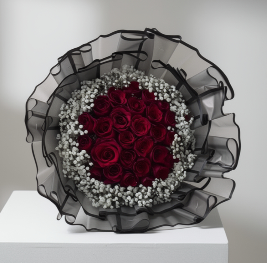 Bouquet of red roses with white flowers in a decorative black and sheer wrap on a white pedestal.